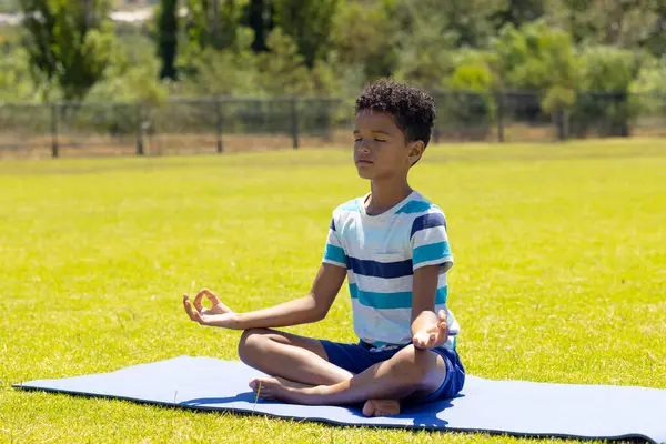 stock image Biracial boy meditates on a yoga mat in a sunny park. He is focused, wearing a striped shirt and shorts, promoting wellness and tranquility.