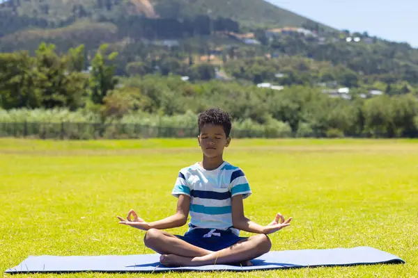 stock image Biracial boy meditates on a yoga mat in a sunny park with copy space. Eyes closed, he exhibits tranquility against a backdrop of green hills and clear skies.