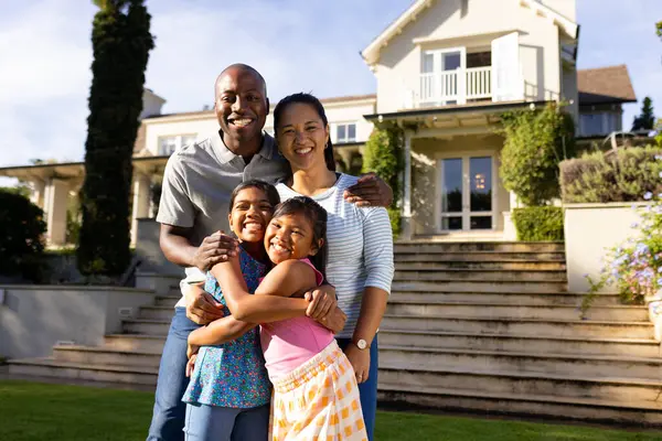 Stock image Outdoors, diverse family of four smiling and embracing in front of their home. The background shows a beautiful, well-maintained house with a lush garden and stone steps, unaltered