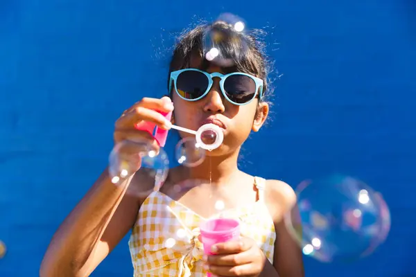 stock image Biracial girl wearing sunglasses and a sundress enjoys blowing bubbles outdoors. She creates a playful atmosphere on a sunny day.
