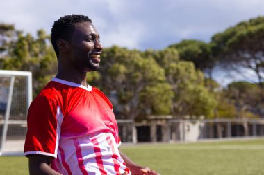 African American male fitness trainer wearing red sports jersey smiling outdoors, copy space. Soccer field with goalpost and trees in background, unaltered clipart