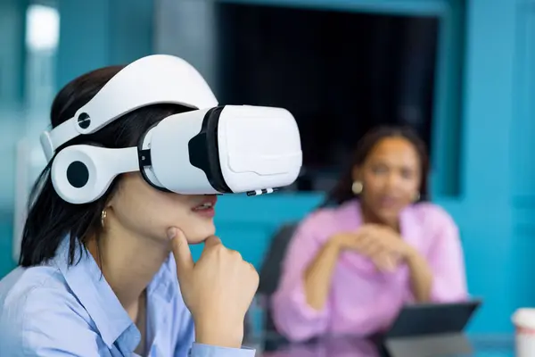 stock image Asian young woman uses VR headset as biracial young woman observes, collaborating on business. They are in modern office with large screen and tablet on table, unaltered