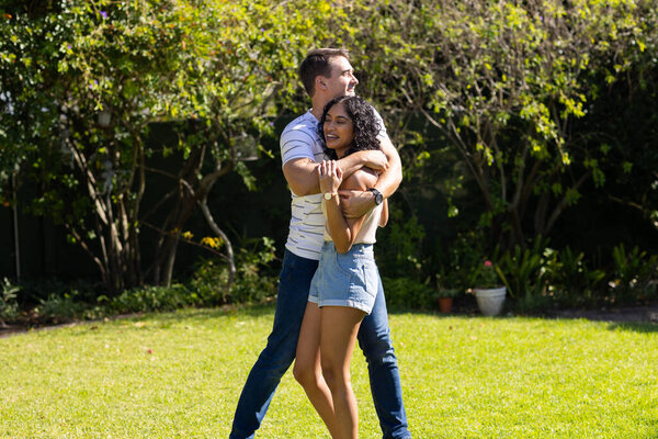 Young Caucasian man hugging young Asian woman from behind, both smiling and happy. They are standing in sunny garden with lush greenery and trees in background, unaltered — Stock Image Young Caucasian Man Hugging Young Asian Woman Both Smiling Happy Stock Photo
