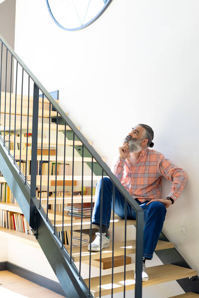 At home, Sitting on stairs, senior man in casual clothes thinking, bookshelf in background. Elderly, contemplation, thoughtful, indoor, unaltered — Stock Image Home Sitting Stairs Senior Man Casual Clothes Thinking Bookshelf Background Stock Picture