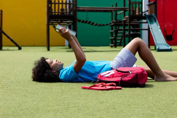 stock image Smiling biracial schoolgirl lying on her back in school playground reading book, with copy space. Education, childhood, elementary school, inclusivity and learning concept.