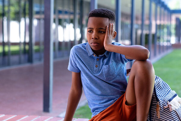 Sad african american schoolboy with schoolbag sitting on wall outside school, with copy space. Education, childhood, inclusivity, elementary school and learning concept. — Stock Image Sad African American Schoolboy Schoolbag Sitting Wall School Copy Space Stock Picture