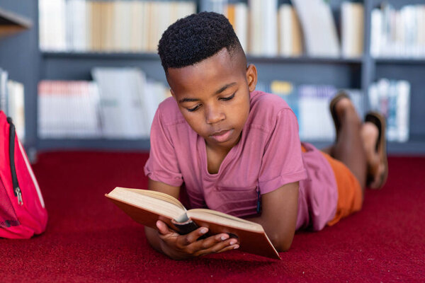 African american schoolboy lying on floor reading book in school library, concentrating, copy space. Education, childhood, elementary school and learning concept. — Stock Image African American Schoolboy Lying Floor Reading Book School Library Concentrating Royalty Free Stock Images