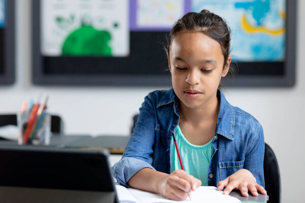 Biracial schoolgirl sitting at desk with tablet, writing in class, concentrating, with copy space. Education, childhood, elementary school, inclusivity and learning concept. — Stock Image Biracial Schoolgirl Sitting Desk Tablet Writing Class Concentrating Copy Space Royalty Free Stock Photos