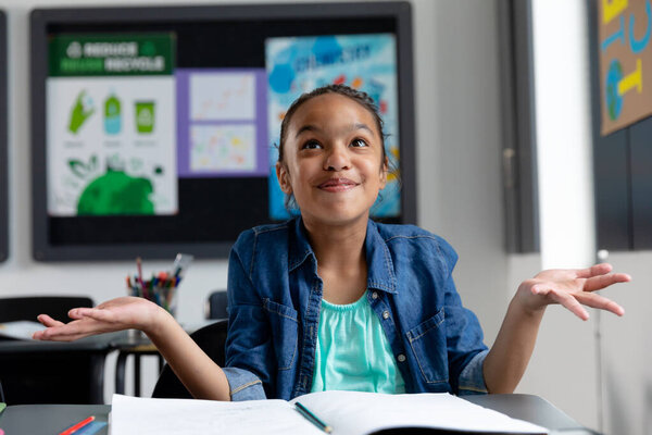 Confused biracial schoolgirl sitting at desk in class, shrugging shoulders, smiling, with copy space. Education, childhood, elementary school, inclusivity and learning concept. — Stock Image Confused Biracial Schoolgirl Sitting Desk Class Shrugging Shoulders Smiling Copy Stock Photo