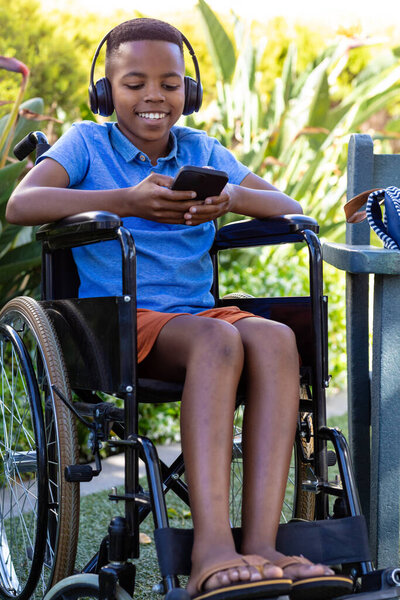 Happy african american schoolboy in wheelchair using headphones and smartphone in garden, copy space. Education, childhood, inclusivity, elementary school and learning concept. — Stock Image Happy African American Schoolboy Wheelchair Using Headphones Smartphone Garden Copy Royalty Free Stock Images