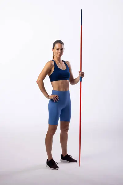 stock image Young Caucasian female athlete poses confidently with a javelin on a white background. Her athletic attire suggests a focus on track and field sports.