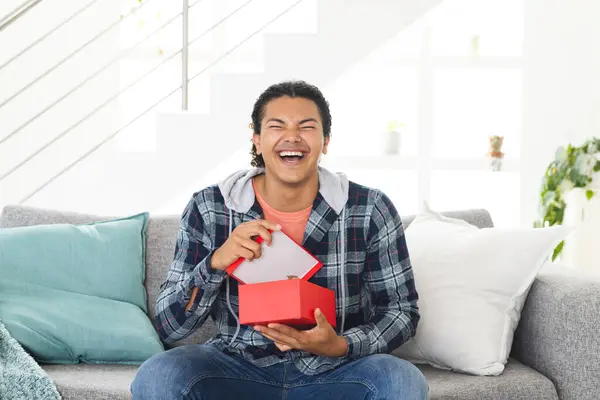 stock image Young biracial man laughing joyfully at home during a video call, holding a gift box. His expression conveys happiness and surprise during a casual celebration.