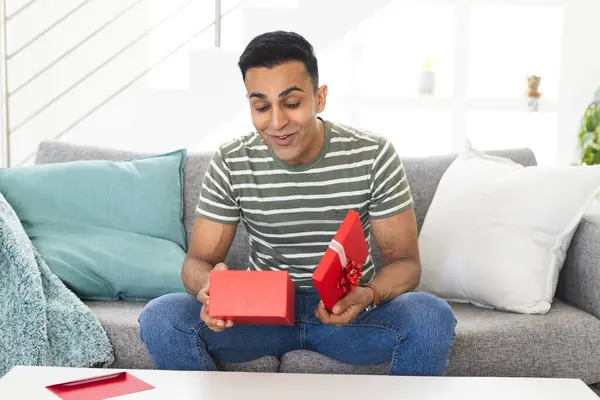 stock image Middle Eastern man opens a gift at home during a video call, with copy space. He exhibits excitement and surprise as he unwraps the present on the couch.