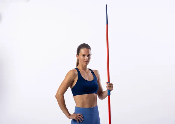 stock image Confident young Caucasian female athlete poses with a javelin on a white background, with copy space. Her athletic attire and posture suggest a professional sports setting.