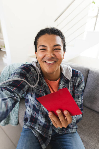 Young biracial man holding a red envelope at home during a video call, with copy space. His cheerful expression suggests a moment of joy or celebration. — Stock Image Young Biracial Man Holding Red Envelope Home Video Call Copy Stock Image