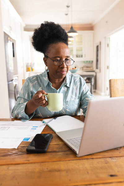 Busy senior african american woman with documents on table using laptop at home. Domestic life, retirement, technology and lifestyle, unaltered. — Stock Image Busy Senior African American Woman Documents Table Using Laptop Home Stock Photo