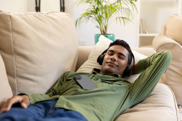 Young biracial man relaxes on a couch at home, with copy space unaltered. He enjoys music with headphones, creating a serene atmosphere. — Stock Image Young Biracial Man Relaxes Couch Home Copy Space Unaltered Enjoys Stock Picture