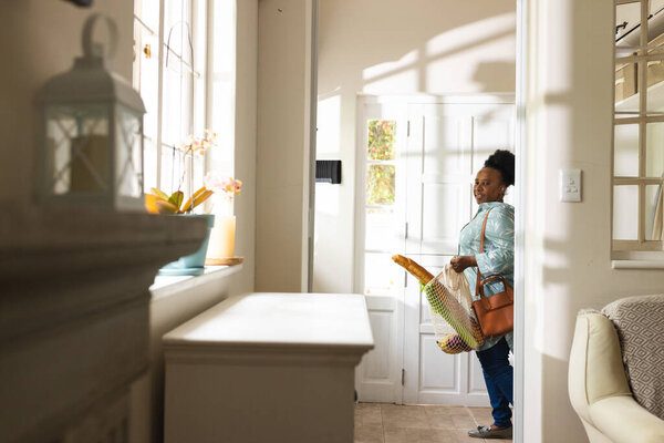 Happy senior african american woman opening door and holding shopping bag at home. Domestic life, retirement and lifestyle, unaltered. — Stock Image Happy Senior African American Woman Opening Door Holding Shopping Bag Stock Photo