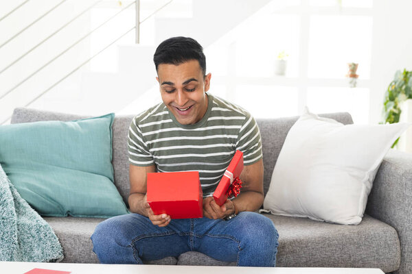 Middle Eastern man looks surprised at a gift during a video call, seated on a couch at home. His joyful expression suggests a special occasion or celebration. — Stock Image Middle Eastern Man Looks Surprised Gift Video Call Seated Couch Stock Image
