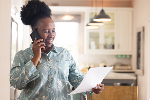 Happy senior african american woman with documents talking on smartphone at home. Domestic life, retirement, technology and lifestyle, unaltered. — Stock Image Happy Senior African American Woman Documents Talking Smartphone Home Domestic Stock Picture