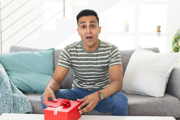 Middle Eastern man sitting on a couch at home during a video call, with copy space. He appears surprised while holding a red gift box, capturing a moment of excitement. — Stock Image Middle Eastern Man Sitting Couch Home Video Call Copy Space Stock Picture