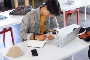 In school, asian teenage boy writing in notebook while using laptop at desk. Education, studying, learning, technology, classroom, academic clipart