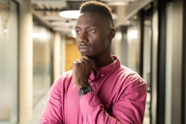 Thinking deeply, african american man in pink shirt standing in modern office hallway. Business, corporate, contemplation, concentration, professional, workplace clipart