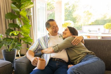 Sitting on couch, couple hugging and smiling while holding coffee mug indoors. love, cozy, happiness, bonding, relaxation, intimacy clipart