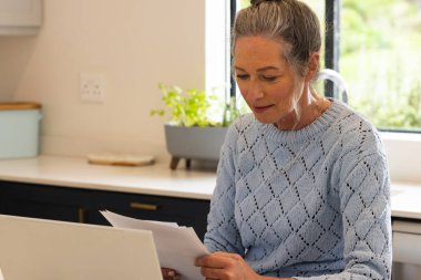 Mature woman reviewing documents at home, focused and thoughtful in kitchen. Home office, paperwork, concentration, lifestyle, mature adult clipart