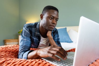 African American man using laptop on bed, focusing on work at home. Remote working, technology, productivity, home office, online, business clipart