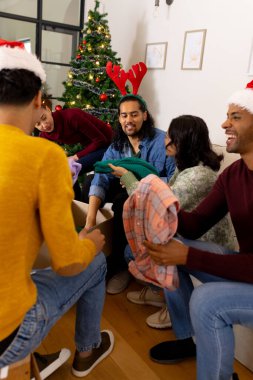 Exchanging gifts by christmas tree, multiracial friends smiling joyfully, at home. celebration, friendship, holiday, winter, gifting, cheerful clipart