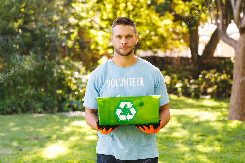 Male volunteer holding recycling bin and wearing orange gloves in sunlit garden promoting recycling. Sustainability, environmental, ecofriendly, conservation, stewardship, community, waste-management