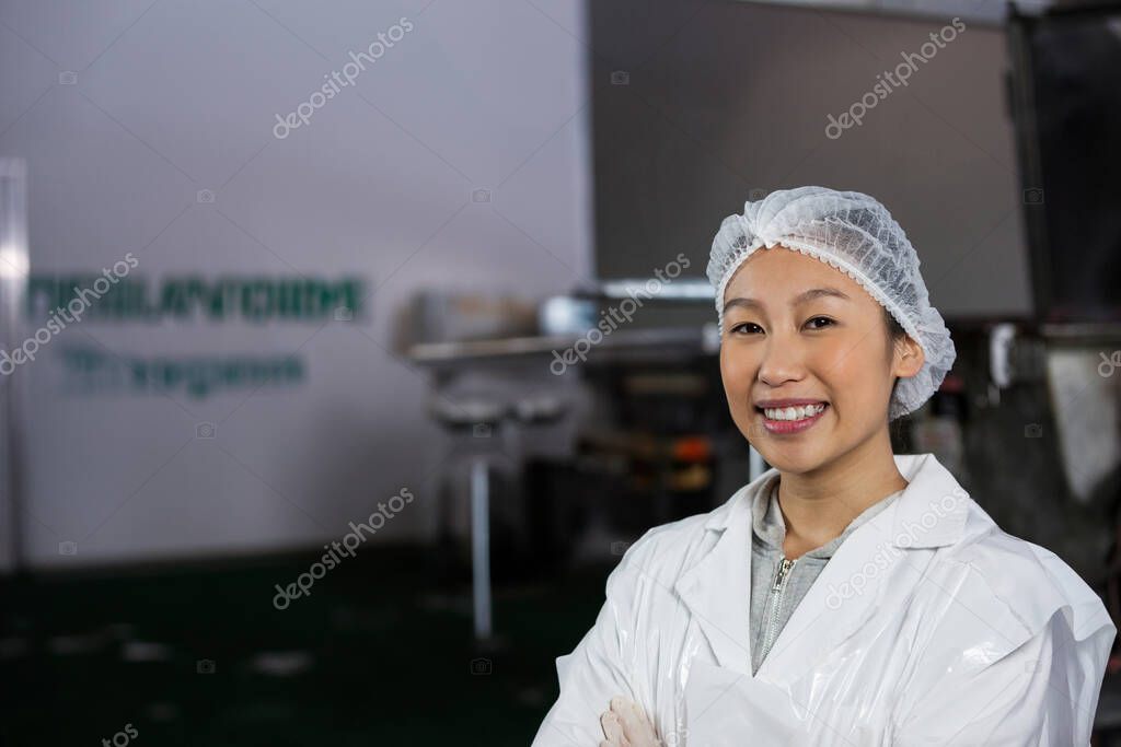 Woman standing, smiling with crossed arms in food processing plant, lab coat, hair net, copy space. Industrial, manufacturing, stainlesssteel, machinery, quality control, inspection, hygiene