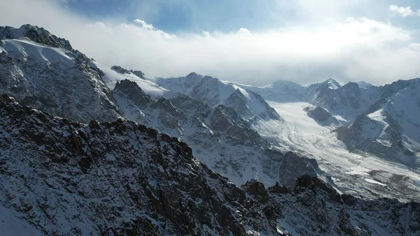 高い山の中で古代の雪の氷河 山の峰々は雪に覆われている 場所には急な崖や大きな岩があります 白い雲と青空 ドローンからの眺め — ストック写真