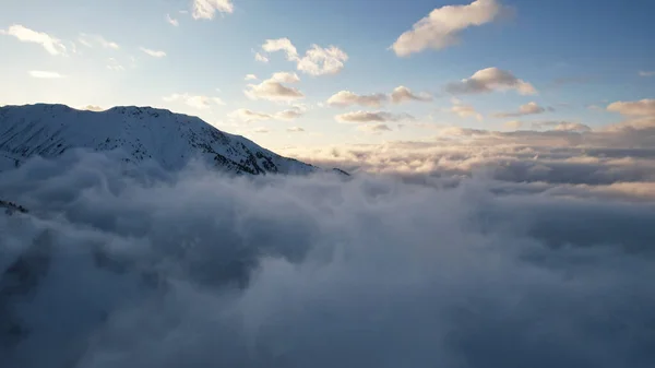 Flying among the ocean of clouds in the mountains. The fog is like big ocean waves crashing against high mountains covered with snow. Spruce trees grow in places. Sunset. Orange rays of the sun