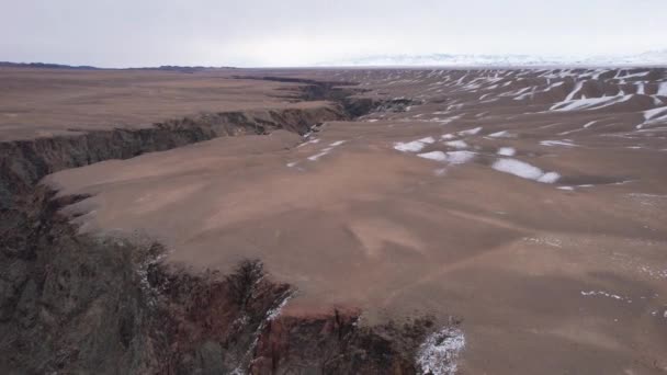 地面上一个巨大的裂缝 俯瞰着雪山 灰蒙蒙的天空布满了云彩 大峡谷在大草原的中央 这条河流过 从无人机上俯瞰 哈萨克斯坦 黑峡谷 — 图库视频影像