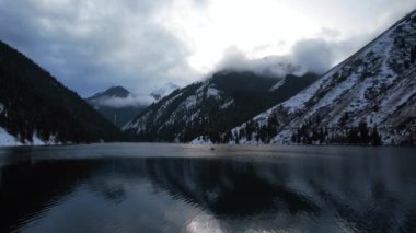 Dağ gölü Kolsai 'nin inanılmaz yüzeyi. Bir ayna gibi su, güneş ışınlarını, beyaz bulutları, karlı dağları ve ormanı yansıtır. İnsanlı tekneler görünür. Üst görünüm
