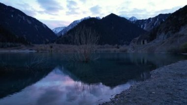 Gün batımında aynalı su ile Issyk Dağı Gölü. Suyun rengi gözlerimizin önünde değişir. Temiz suda ağaçlar var. Karlı dağlar ve yeşil tepeler görülebilir. Bulutlar yansıtılır