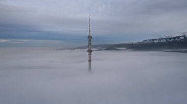 TV kulesi gün batımında bulutlardan dışarı bakıyor. İHA 'nın çift katmanlı bulutlar üzerindeki görüntüsü. Bulutların sarı-mavi tonu. Bulutlardaki gölgeler. Uzaktan yüksek dağlar görülebilir. Kok Tobe