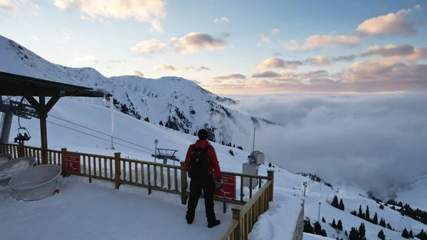A guy with a backpack is standing on an observation deck in the snowy mountains. In distance, snow-white clouds and fog floating along gorge. A pink sunset is visible. An icicle hangs from the roof
