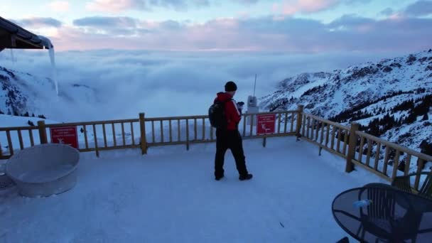 雪の山の展望台にはリュックを背負った男が立っている 遠くには 雪の白い雲と霧の峡谷に沿って浮動小数点 ピンクの夕日が見える 屋根からつららが垂れている — ストック動画