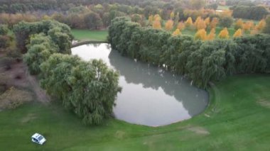 Gün batımında söğüt ağacı ve gölet manzaralı yeşil bir golf sahası. Göletteki su gri, ağaçları ve güneşi yansıtıyor. Kuşlar yüzüyor. Güneş puslu bir havada. Sarı-kırmızı yapraklar. Sonbahar