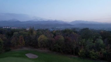 Karlı tepeler manzaralı sonbahar golf parkı. Altın ağaçların, sarı-kırmızı yaprakların ve yeşil otların hava manzarası. Evler ve dağlar uzaktan görülebilir. Gün batımı. Kazakistan