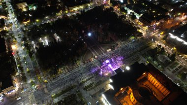 Gece şehrindeki büyük bir konserin insansız hava aracı görüntüsü. Bir sürü insan, ışık huzmeleri. Işıklı büyük bir bina. Arabalı sokaklar var, insanlarla dolu yaya sokakları ve parkı var. Almaty