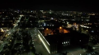 Gece şehrinde büyük bir konser. Kalabalığın insansız görüntüsü, ışınları ve ışık huzmeleri. Işıklı büyük bir bina. Arabalı yollar, yayalı sokaklar ve etrafta bir park var.