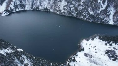 Kolsai Dağı Gölü, kış ormanında. Bulutların, kozalaklı ağaçların, aynadan yansıyan suyun, kardaki tepelerin ve dağların drone görüntüsü. Sarı günbatımı. Tekneler bazı yerlerde yüzer. Kazakistan, Almaty