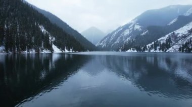 Kolsai Dağı Gölü, kış ormanında. Ahşap bir iskele, beyaz bulutlar, kozalaklı ağaçlar, ayna gibi su, tepeler ve kardaki dağlar. Sarı günbatımı. Tekneler bazı yerlerde yüzer..