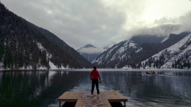 İskelede duran adam dağ gölüne hayran. Suyun, karlı dağları, yeşil ormanı, bulutları ve sarı bir günbatımını yansıtan siyah aynalı bir görüntüsü vardır. Kolsai Gölü