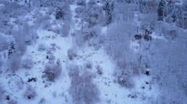 Kış ormanlarındaki dağlarda kablo tv arabası. Kulübeler, kar ormanları ve sis boyunca gondol yolu boyunca ilerliyorlar. Dağları beyaz bulutlar kapladı. İHA 'nın üst görüntüsü. Medeo, Almaty