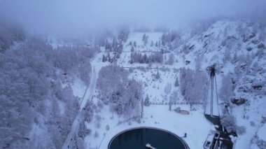 Almaty 'de kozalaklı ağaçlarla kaplı karlı dağlar. Dağ suyu için bir rezervuar. Medeo Barajı ve sağlık merdivenleri. Her şey bulutlar ve karla kaplı. İnsanlar ve teleferik görünüyor.